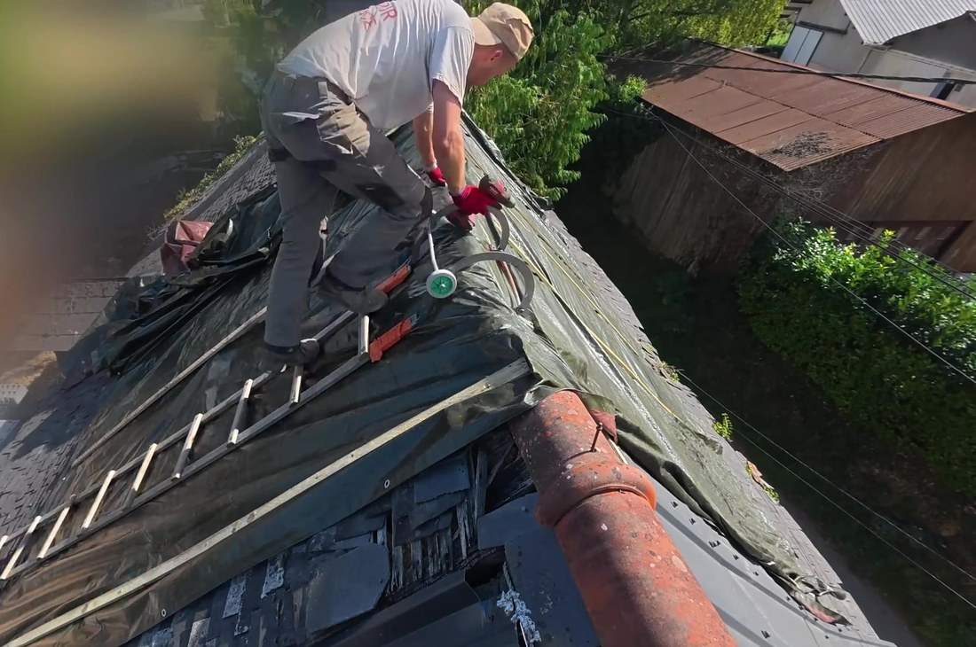 Couvreur fixant une bâche verte sur un rampant en ardoise, échelle de toit posée sur la couverture