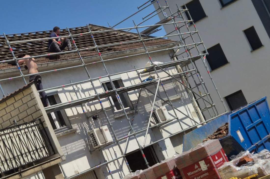 Chantier de charpente sur une maison sous échafaudage, benne et palettes de matériaux au pied de la façade