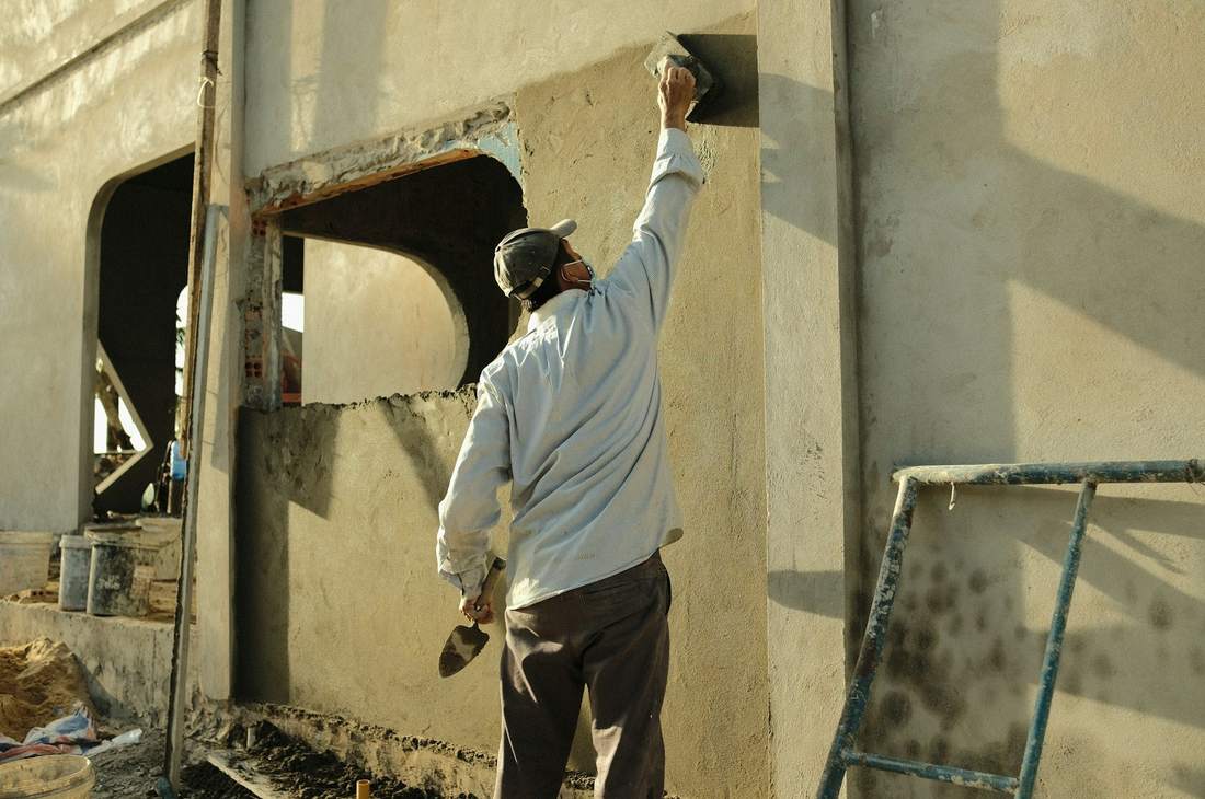 Ouvrier étalant un enduit sur un mur à la taloche, truelle dans l'autre main